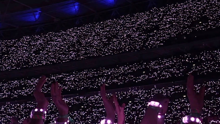 Auf dem Bild ist ein Stadion mit vielen Menschen zu sehen. Da es sonst dunkel ist, sind im Hintergrund viele Lichter zu sehen, die die Masse an Menschen zeigen. Im Vordergrund sind Arm mit leuchtenden Bändern zu sehen, die über der Menschenmasse schweben. Es handelt sich von einer Aufnahme auf einem Konzert.