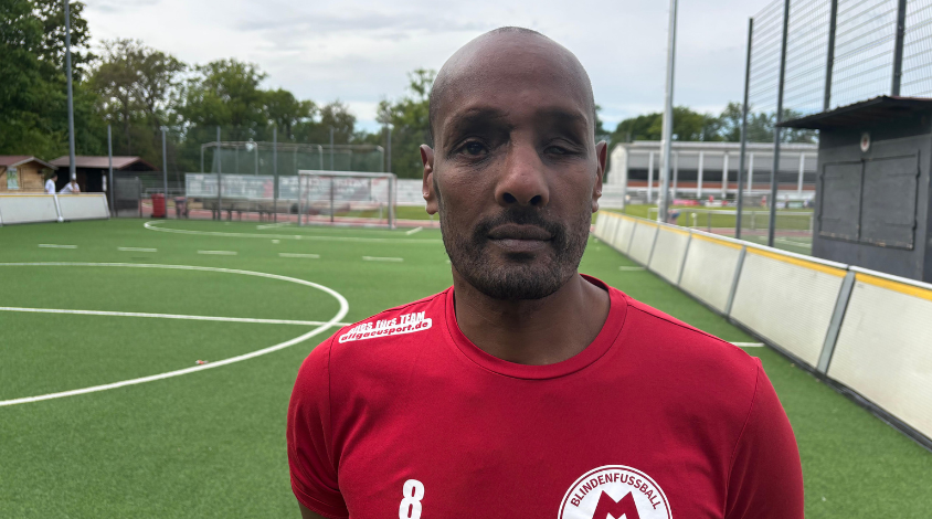Auf dem Bild ist im Fokus ein dunkelhäutiger Mann mit einem roten Trikot zu sehen. Er steht auf der rechten Seite und schaut Richtung Kamera. Im Hintergrund sind man ein kleines Fußballfeld. Auf der rechten Seite ist hinter ihm eine Bande zu sehen, welche die Seitenränder bilden. 