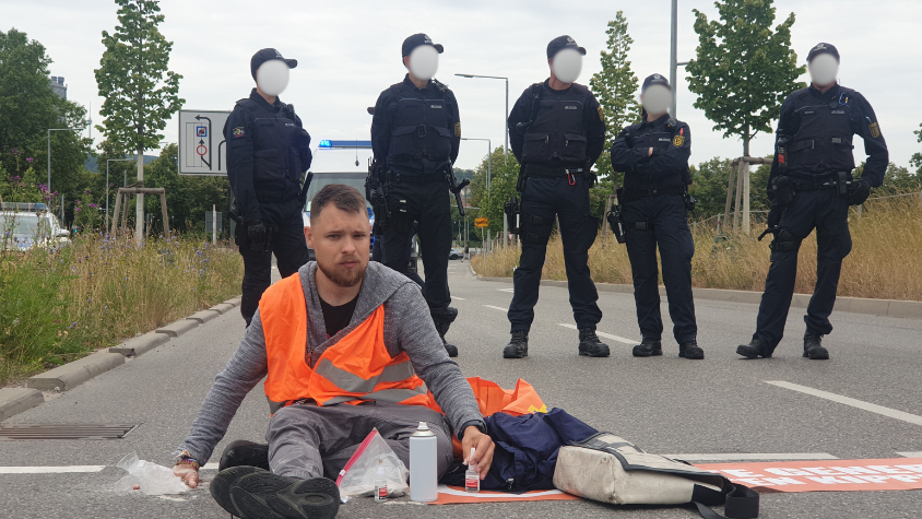 Der Klimaaktivist Moritz Riedacher klebt sich auf der Straße fest. Hinter ihm stehen aufgereiht Polizisten.