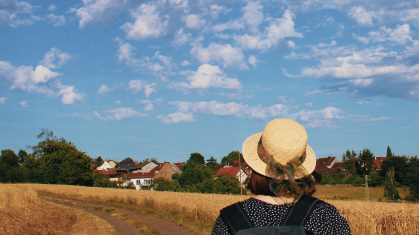 Eine Frau steht auf einem Feldweg und schaut auf ein Dorf in der Ferne.