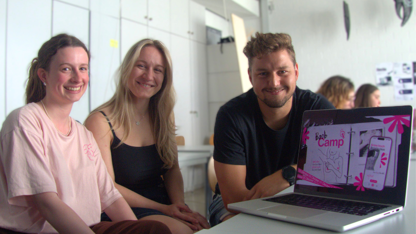 Drei Mitglieder des Boob-Camp-Teams lächeln hinter einem aufgestellten Laptop, auf dem der Titel ihres Projekts zu sehen ist, in die Kamera