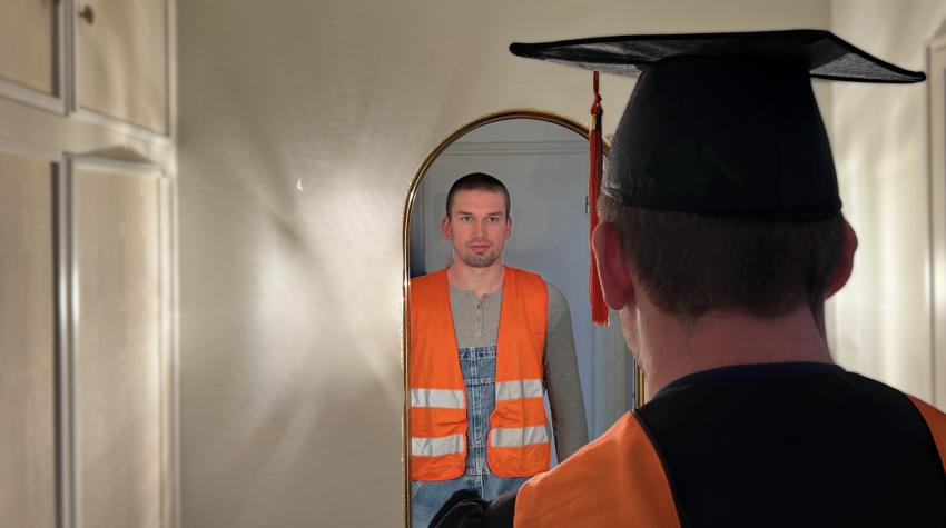 Ein Mann mit Absolventenhut blickt in einen Spiegel und sieht sich dort in Arbeitskleidung mit Warnweste und Blaumann.