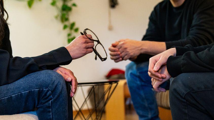 Eine Damenhand, die eine Brille hält und gegenüber einem Pärchen sitzt.