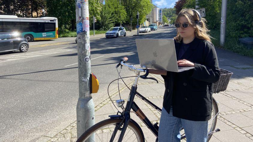 Eine Frau steht an einer Ampel. In einer Hand hält sie einen Laptop, in der anderen ein Fahrrad. 