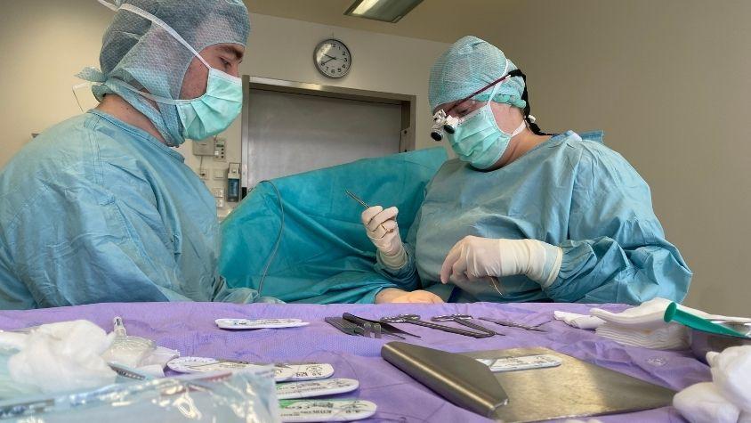 Zwei Chirurg*innen sitzen in sterilen Kitteln an einem OP-Tisch und operieren an einer Hand, im Vordergrund steht ein Tisch mit Instrumenten