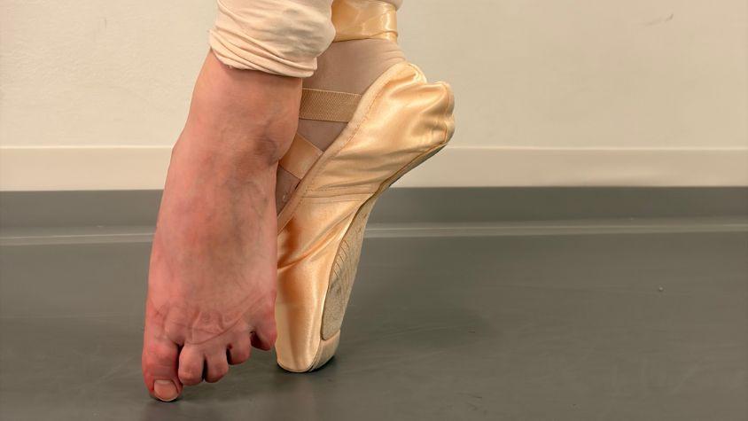 Eine Balletttänzerin zeigt die fünfte Position auf Spitze. Die Füße stehen eng voreinander, die Ferse eines Fußes berührt die Zehen des anderen, beide Beine und Füße sind auswärts gedreht. Ein Fuß ist nackt, am anderen ist ein Spitzenschuh.