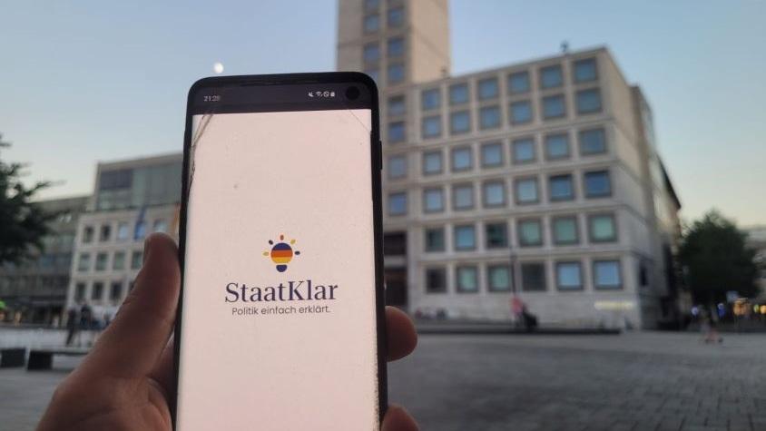 Bild eines Handys vor dem Stuttgarter Rathaus. Auf dem Handy ist das "StaatKlar"-Logo zu sehen.