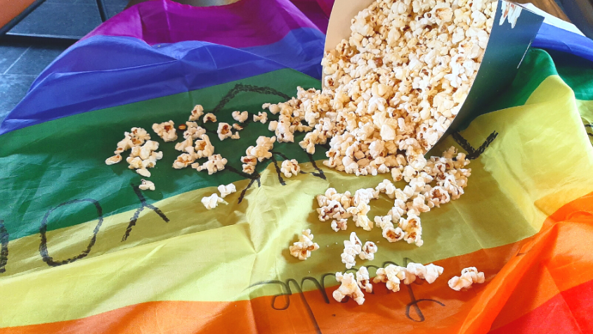 Auf dem Bild sieht man eine Regenbogenflagge, die auf einem Tisch liegt. Rechts oben im Bild liegt ein umgeworfener Popcornbecher. Das Popcorn fällt aus dem Becher über die Flagge.