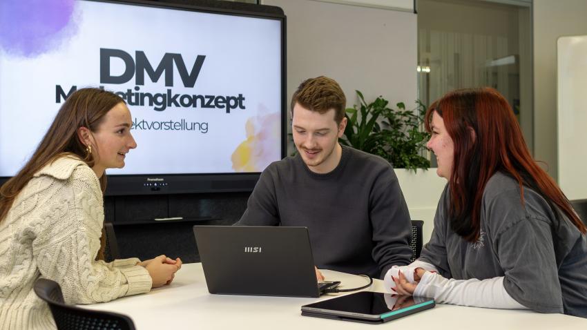 Studierende der HdM sitzen an einem Tisch, diskutieren angeregt und planen ein Marketing-Projekt; im Hintergrund ein Bildschirm mit „DMV-Marketing-Konzept“.