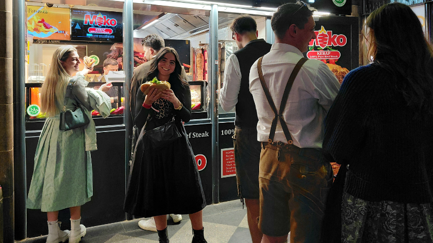 In der Mitte des Bilds ist eine Frau in einem Dirndl vor einem Dönerladen zu sehen, die einen Döner in ihren Händen hält. Auf der rechten Seite des Bilds sieht man eine Warteschlange. Auf der linken Seite nimmt gerade eine Frau einen Döner entgegen. Alles Personen auf dem Bild sind mit Dirndl oder Lederhose gekleidet. 