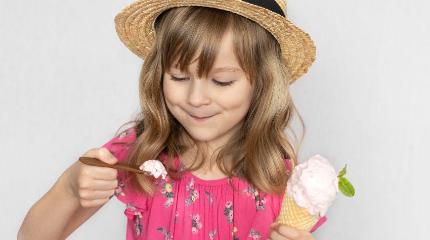Kleines Mädchen, welches in der linken Hand einen essbaren Löffel mit Eis hält und in der rechten eine Eiswaffel.