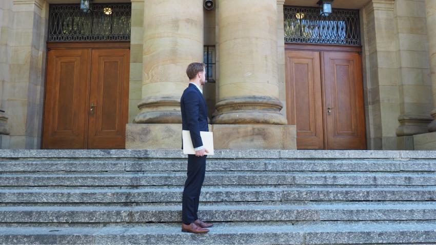 Ein Mann im Anzug/Business Kleidung steht auf einer Treppe vor zwei Türen und dreht seinen Kopf unentschlossen in die Richtung der Türen, als überlege er durch welche er gehen soll.