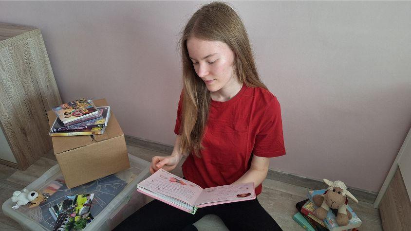 Mädchen sitzt auf dem Boden und blättert ein Buch durch. Um sie herum Kisten voll mit Kindheitserinnerungen.