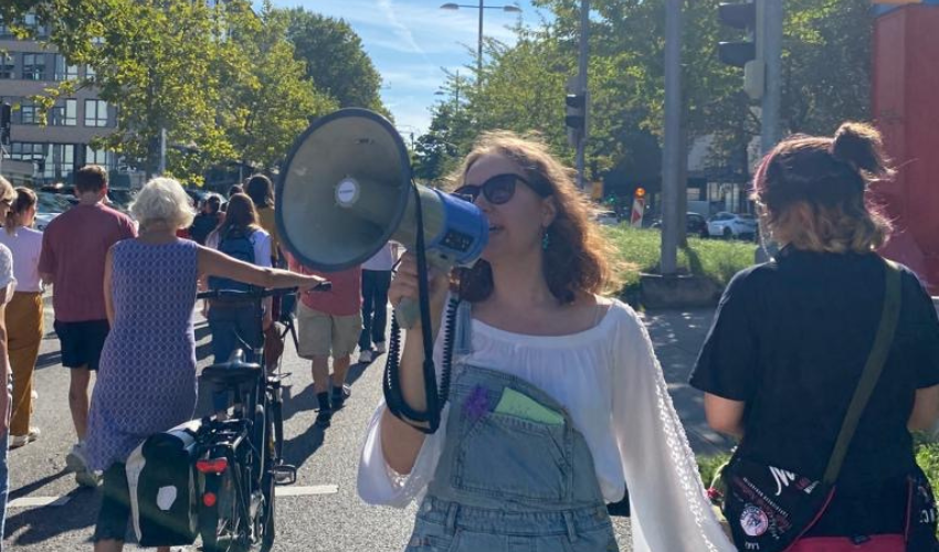 Eine junge Frau mit Sonnenbrille und Latzhose ruft in ein Megaphon. Um sie herum laufen Menschen im Demonstrationszug.