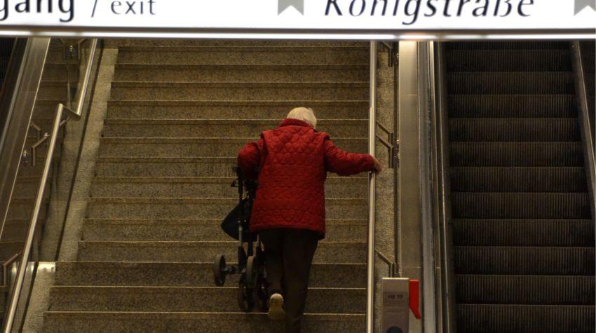 Eine ältere Frau in einem roten Mantel läuft die Treppen einer U-Bahnstation hoch Richtung Königsstraße. Ihren Rollator trägt sie dabei in der Hand. Neben ihr befindet sich eine Rolltreppe.
