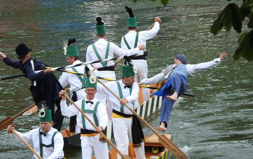 Auf einem Fluss fahren mehre Personen in weißen, traditionellen Gewändern auf jeweils zwei kleinen Boot. Zusätzlich dazu stehen auf jedem Boot ein weitere Mann in anderen Outfits, die jeweils eine Stange in der Hand halten. Beide Männer kippen nach hinten und scheinen in den Fluss zu fallen.