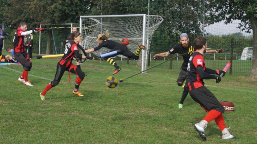 Mehere Spieler spingen auf einer grünen Wiese mit einem Fußballtor im Hintergrund verschiedene Sportgeräte um sich. Zu diesen Sportgeräten zählen Bälle an Seilen und gepolsterte Stäbe. Die Spielerinnen und Spieler tragen jeweils rot-schwarze oder gelb-schwarze Trikots.