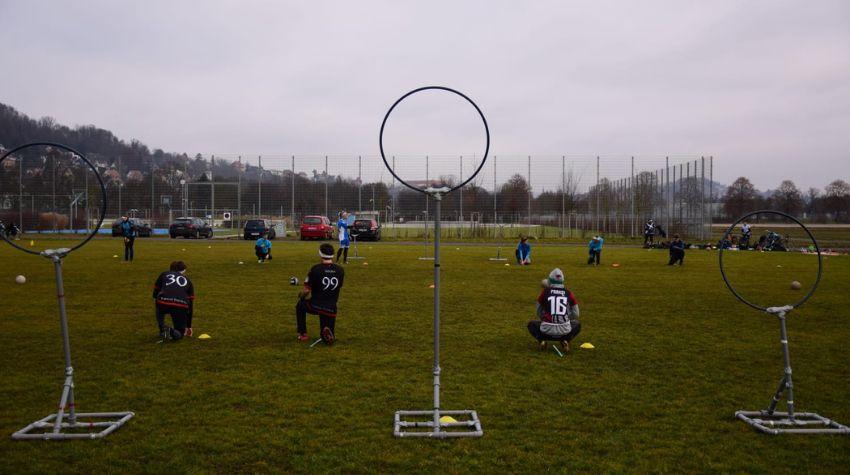 Auf einer Wiese stehen drei Stäbe mit großen Ringen am oberen Ende, wobei der Stab in der Mitte der größte ist. Vor diesen Ringen knien drei Spieler mit schwarzen Trikots.