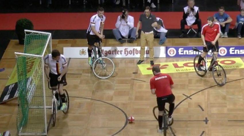 Mehrere Spieler auf Fahrrädern stehen in einer Sporthalle. Auf der linken Seite steht ein Tor, kurz davor liegt ein kleiner rot-weißer Ball. Das Spielfeld ist durch Werbebanner begrenzt, hinter diesen Banner sitzen mehrere Fotografen.