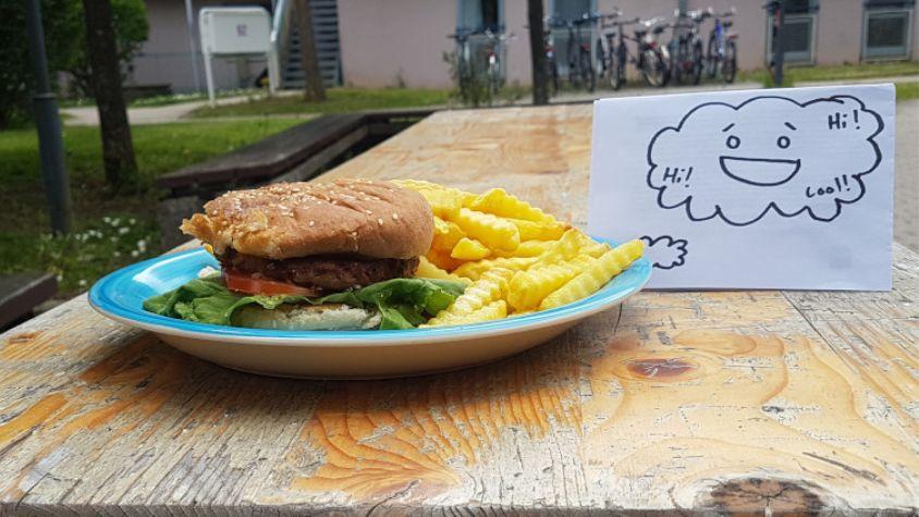 Auf einem blauen Teller liegt ein Burger mit Pommes. Daneben ist ein Blatt Papier aufgestellt, auf dem eine Gedankenwolke mit einem lachenden Gesicht und verschiedenen Worten gezeichnet. Alles steht auf einem Biertisch in einem Innenhof.
