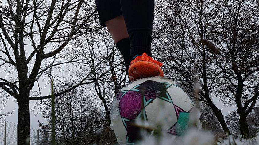 Eine Frau steht mit orangefarbenen Fußballschuhen mit einem Bein auf einem Fußball, der auf einer schneebedeckten Wiese liegt.