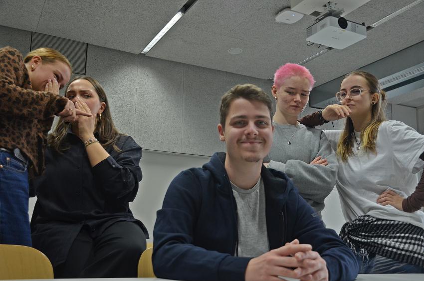 Ein Mann schaut lächelt künstlich in die Kamera. Im Hintergrund blickt eine Gruppe auf ihn.