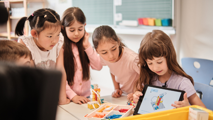 Kids are sitting in a classroom around the table looking at little pieces to build a robot out of lego.