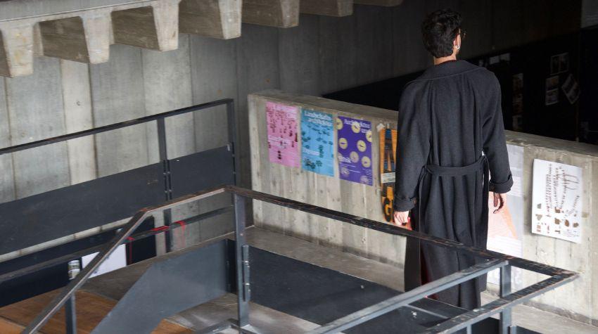 Beton-Treppenhaus ist zu sehen. An der rechten Ecke des Treppenhauses geht Kaan mit dem Rücken zur Kamera gewendet den gang entlang.