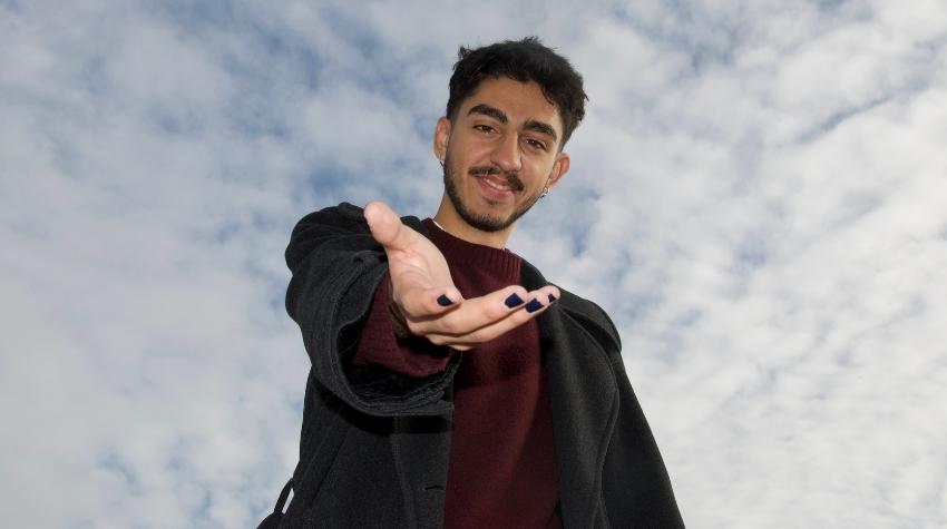 Kaan steht vor einem wolkigen, blauen Himmel und streckt mit einem Lächeln sein Hand in die Kamera.