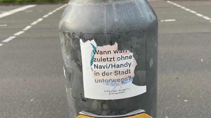 Auf einem abgeblätterten Sticker an einem Ampelpfosten steht "Wann wart ihr zuletzt ohne Handy/ Navi in der Stadt unterwegs?