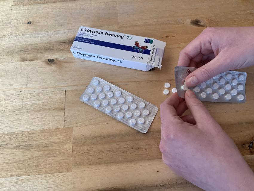 Rechts befinden sich zwei Hände, die Tabletten aus ihrer Verpackung holen. Auf dem Tisch befindet sich zudem die große Verpackung zu den Hormontabletten L-Thyroxin. 
