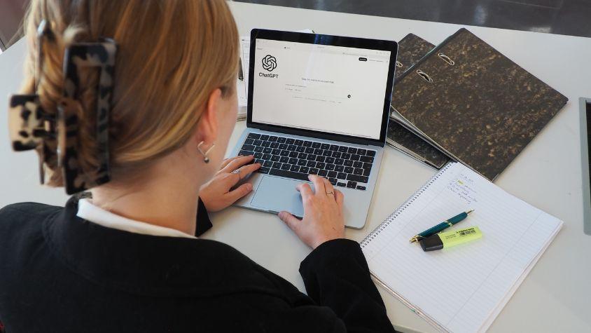 Eine junge Frau in Arbeitskleidung sitzt an einem Tisch. Auf dem Tisch liegen Arbeitsmaterialien wie Ordner und ein Block mit Stiften. Die Frau ist jedoch konzentriert auf ihren Laptop, der vor ihr steht, auf dem sie ChatGPT geöffnet hat.