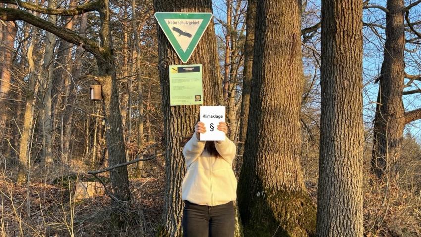 Person steht im Wald und hält ein Buch auf dem „Klimaklage“ steht