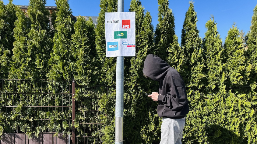 Jugendlicher mit Kapuze geht an Wahlplakat mit Logos etablierter Parteien vorbei und schaut auf sein Smartphone
