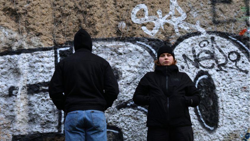 Zwei Menschen stehen vor einer Mauer, einer mit dem Rücken zur Mauer, der andere sieht die Mauer an.