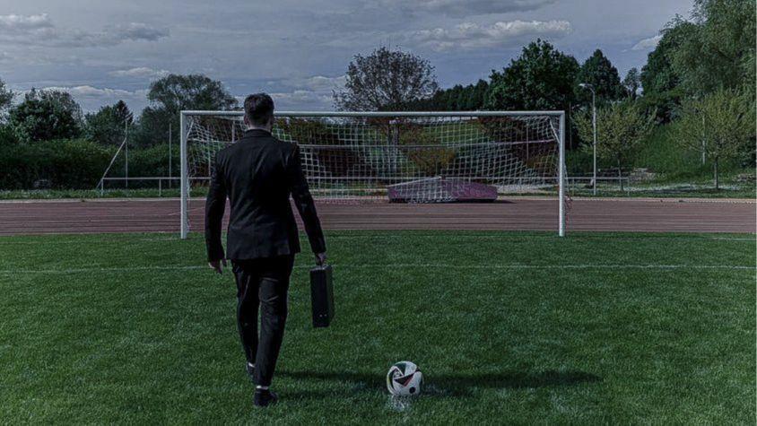 Ein Mann im Anzug mit einem Aktenkoffer in der Hand läuft im Strafraum eines Fußballfelds am Ball vorbei, der auf dem Elfmeterpunkt liegt. Im Hintergrund steht ein Fußballtor.