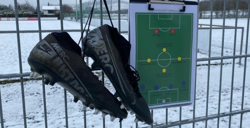 Ein Klemmbrett hängt an einem Gitterzaun, daneben hängen Fußballschuhe und dahinter sieht man einen verschneiten Fußballplatz. 