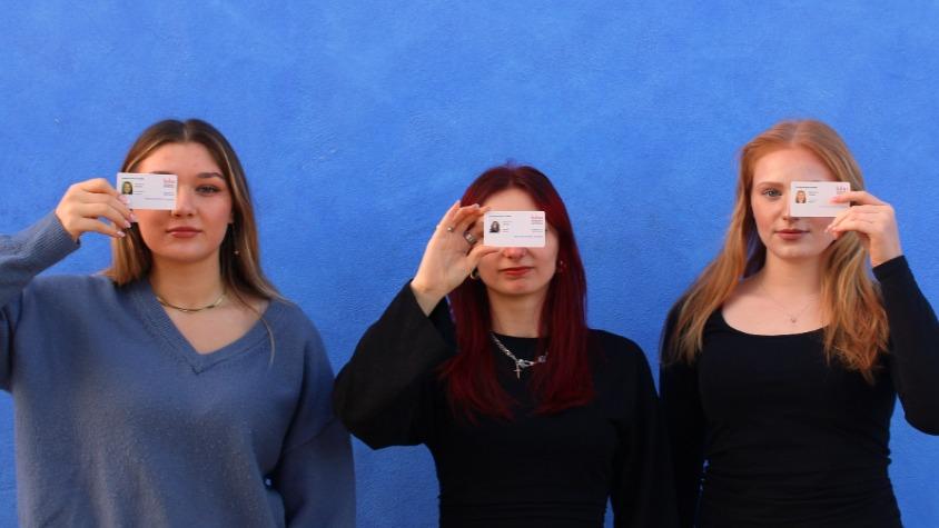 Drei Studentinnen vor einer blauen Wand, die mit ihrem Studentenausweis ihr Gesicht halb verdecken