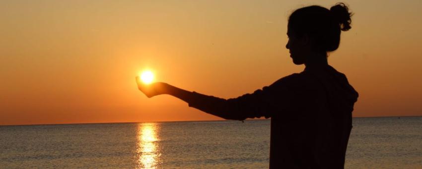 Eine junge Frau hält am Strand ihre Hand der Sonne entgegen. | Bild: Kim Romagnoli