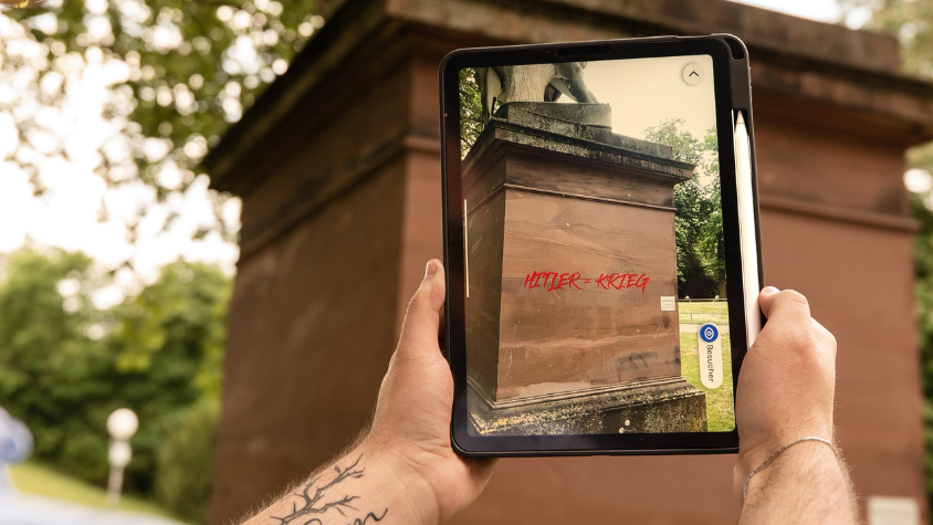 iPad filmt auf den Sockel einer Statue. Durch Augmented Reality wird eine Pinselei mit dem Text "Hitler=Krieg" von Hans Gasparitsch (1918-2002) sichtbar.