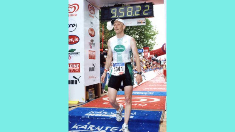 Stefan überquert die Ziellinie des Ironman 2005. Über ihm ist eine Stoppuhr, die 9.58.22 Stunden anzeigt. 