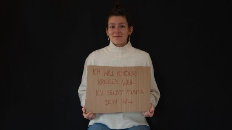 Es ist eine junge Frau mit einem Schild zu sehen. Auf dem Schild steht: Ich will Kinderkriegen, weil ich selbst Mama sein möchte. 