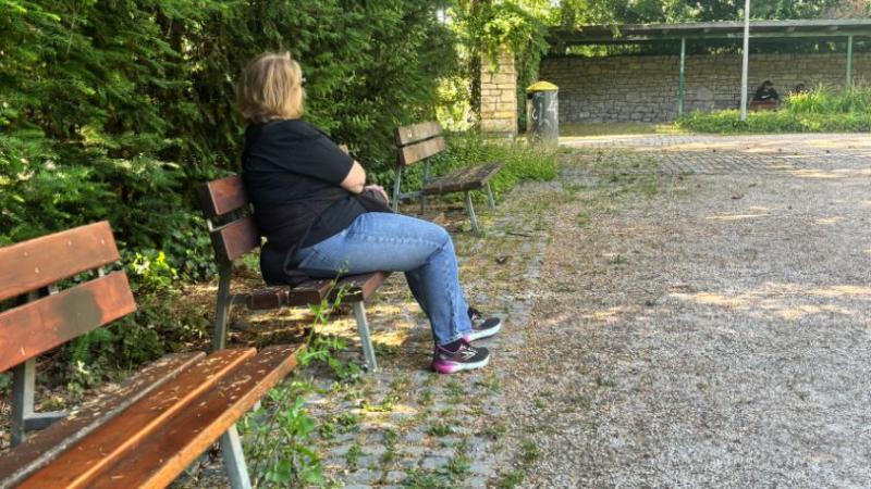 Zu sehen ist eine Frau, die auf einer Bank im Schatten im Kurpark sitzt. Im Hintergrund sitzen außerdem zwei Menschen auf einer überdachten Bank.