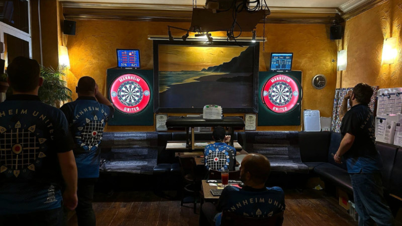 Mehrere Dartsspieler stehen vor zwei beleuchtetet Dartsscheiben. Zwei zählen sitzend die geworfenen Punkte