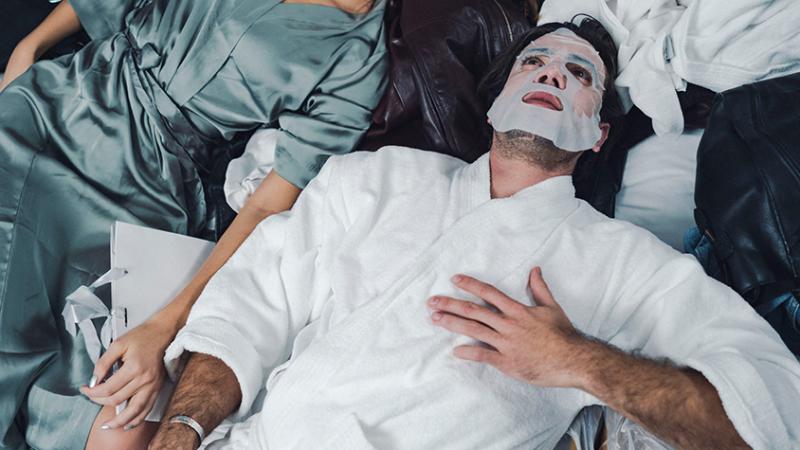 Ein Mann im Bademantel trägt eine Tuchmaske und liegt mit einer Frau in einem Bett.