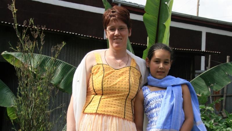 Eine Frau in einem gelbem traditionellem äthiopischen Festkleid hat den Arm um ein Mädchen in einem blauem traditionellem äthiopischem Festkleid gelegt.