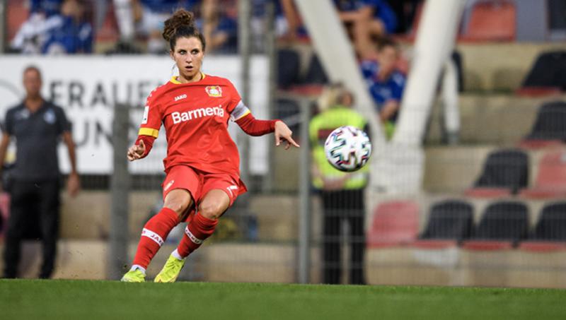Die Fußballerin Jessica Wich von Bayer 04 Leverkusen trägt ein rotes Fußball-Trikot mit neongelben Schuhen und schießt einen Ball, der sich in der Mitte des Bildes verbindet.