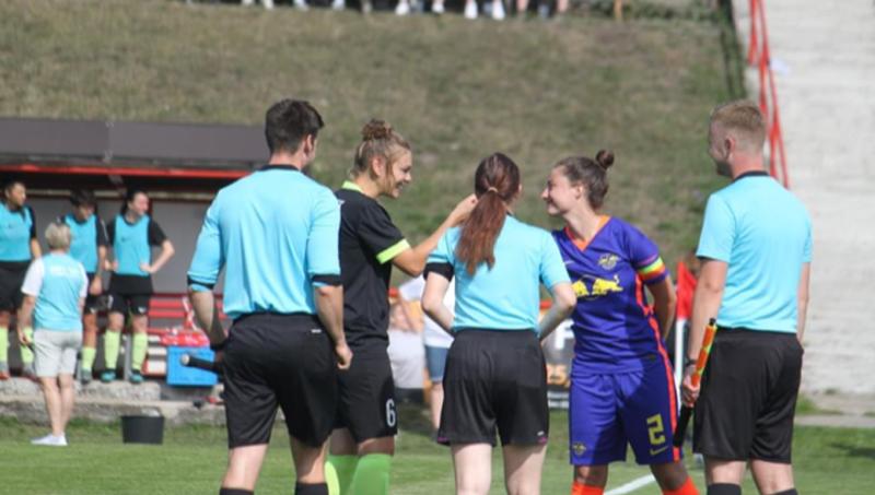 Die Fußballerin Katharina Freitag trägt ein schwarzes Trikot und steht hinter einem Schiedsrichtergespann in blauen Trikots. Sie lächelt und macht einen Handschlag mit einer anderen Fußballspielerin in einem lilanen Trikot.