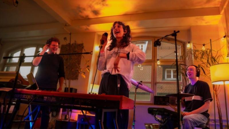 Jasmin steht auf der Bühne mit einem Mikrofon in der Hand. Vor ihr ist ein Keyboard aufgebaut. Im Hintergrund sind ihre Kommilitonen zu sehen. Einer ebenfalls am singen, der zweite am Schlagzeug.