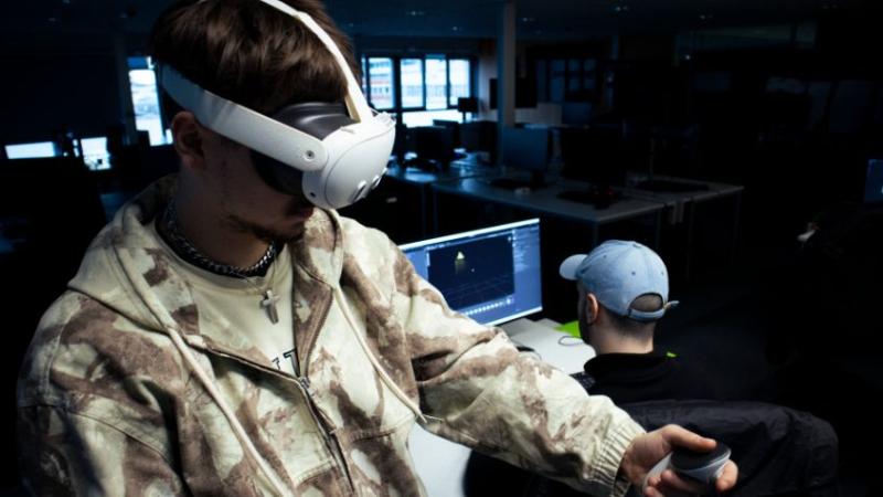 Ein Mann trägt ein VR-Headset. Ein anderer sitzt am Schreibtisch.